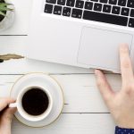 Una pantalla con una web moderna abierta, y una mano tomando notas con una taza de café al lado.