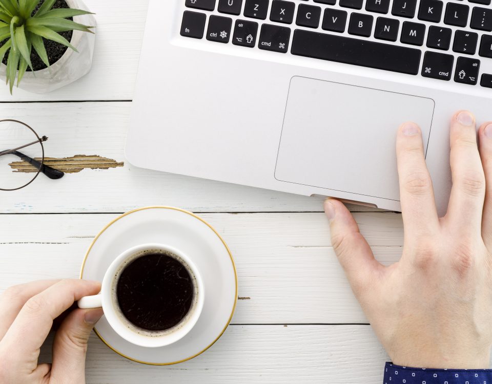 Una pantalla con una web moderna abierta, y una mano tomando notas con una taza de café al lado.